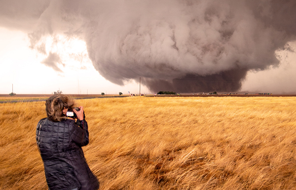 A person in a dark jacket takes a photo of a large, rotating supercell storm over a golden field.