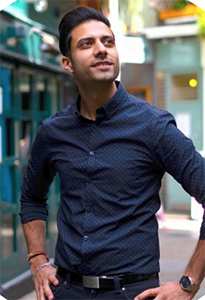 A man wearing a dark blue patterned button-down shirt and black pants stands outdoors, looking upward.
