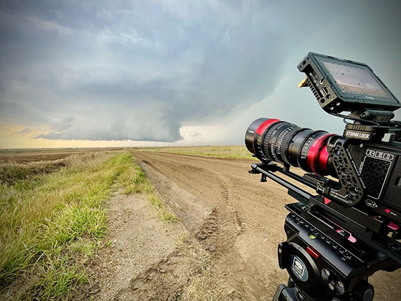 A professional cinema camera in the foreground filming a dark tornado over a grassy field under a stormy sky.
