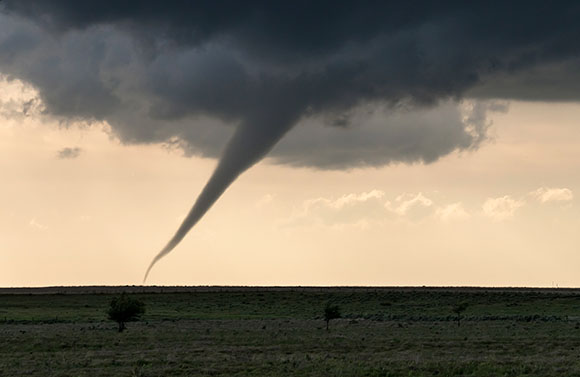 A dark tornado funnel extends from heavy storm clouds to a flat, grassy horizon under a pale sky.