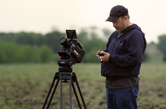 A man in a dark hoodie looks at a device next to a professional RED cinema camera on a tripod in a field.