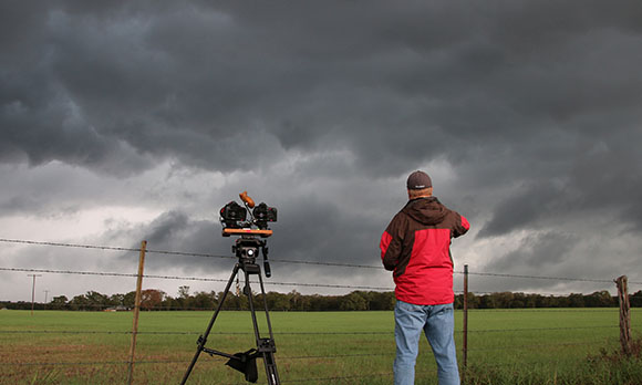A person in a red jacket films dark, heavy storm clouds over a green field behind a barbed wire fence.