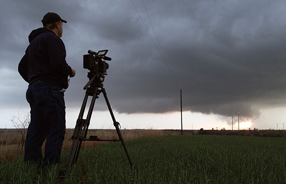 A man stands in a field with a camera on a tripod, filming dark storm clouds approaching a horizon with wind turbines.