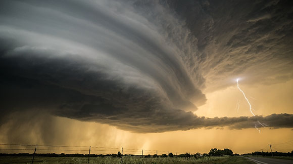 A large, swirling supercell storm cloud hangs over a field as a bolt of lightning strikes the horizon.
