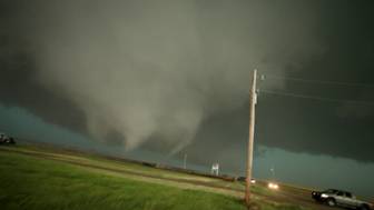 A large, dark tornado descends from a heavy storm cloud toward a grassy field and road.