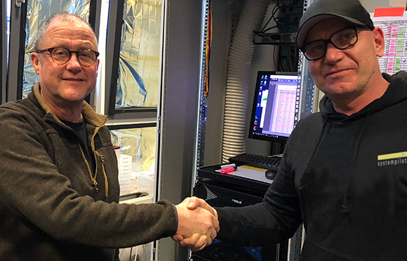 Two men wearing glasses shake hands in a room filled with server equipment and computer monitors.