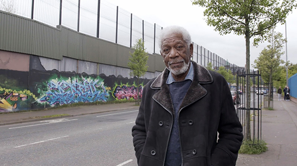 An older man with white hair and a beard wearing a black coat stands on a city sidewalk.