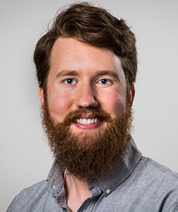 A smiling man with wavy brown hair and a full reddish-brown beard wearing a grey button-down shirt.
