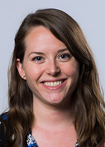 A smiling woman with long brown hair and blue eyes looking toward the camera against a light background.