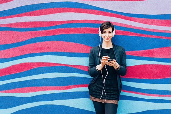 A woman in a dark cardigan uses a smartphone while wearing white headphones against a blue and pink wavy wall.