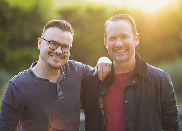 Two smiling men standing close together with arms around each other in a sunlit outdoor setting.