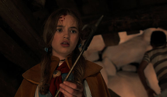 A young girl with a wound on her forehead holds a metal trident-like tool in a dimly lit room.
