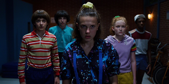 Five teenagers stand in a dark room, looking forward with intense, serious facial expressions.