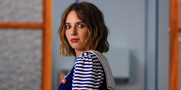 A woman with wavy blonde hair and freckles looks back over her shoulder while wearing a blue and white striped top.