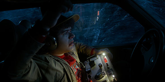 A young man in a hat and headphones looks startled while holding a control device inside a dark car.