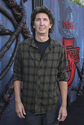 A man with wavy brown hair wearing a dark green plaid shirt stands before a dark movie poster.