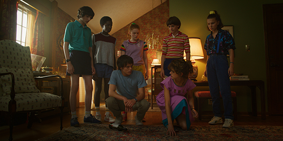 Six teenagers stand and crouch in a living room, looking down with serious expressions.
