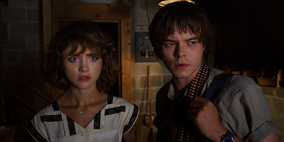 A young woman with curly hair and a young man with dark hair look ahead with startled expressions in low light.
