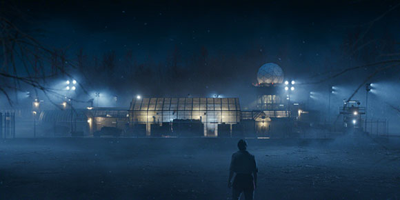 A person stands in a dark, foggy field facing a lit industrial facility with a large geodesic dome.