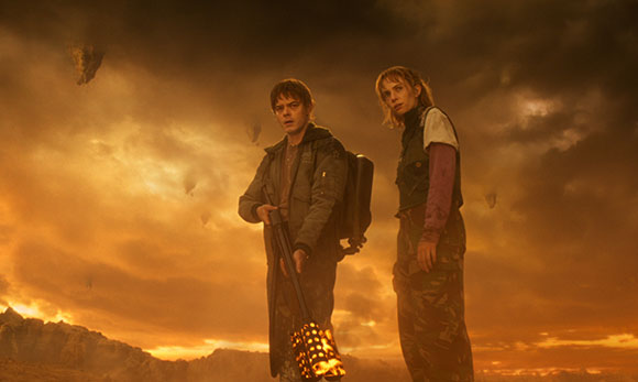 A man holding a glowing staff and a woman stand against a dark, cloudy orange sunset sky.