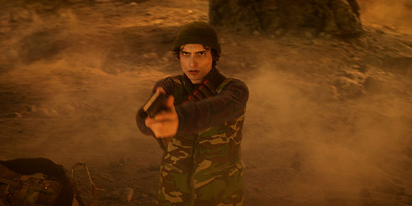 A young man in a dark beanie and camouflage jacket points a handgun forward amidst swirling dust.