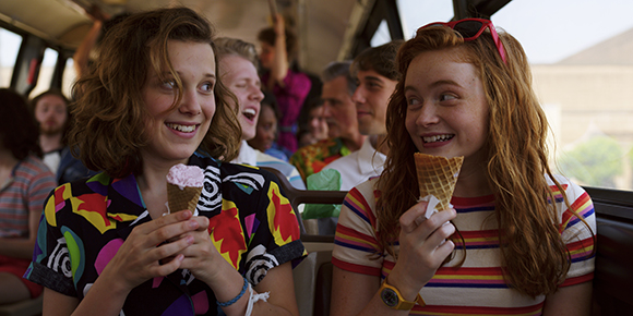 Two teenage girls smile and hold ice cream cones while sitting on a bus with other passengers.