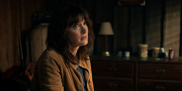 A woman with dark hair and bangs wearing a brown jacket looks up with a worried expression in a dimly lit room.