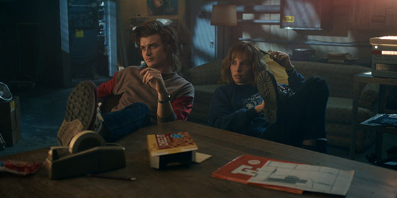 Two teenagers relax on a couch and floor in a cluttered, dimly lit living room.