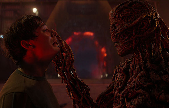 A textured, reddish-brown creature reaches out to touch a man's chin in a dimly lit setting.