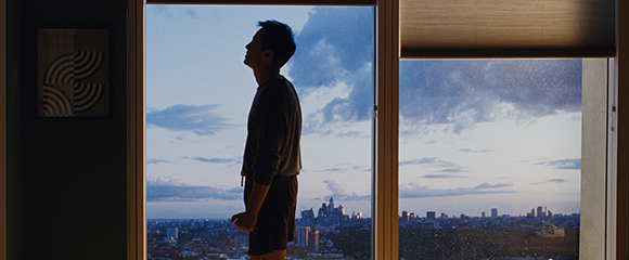 Silhouette of a man looking up through a large window at a city skyline under a cloudy twilight sky.