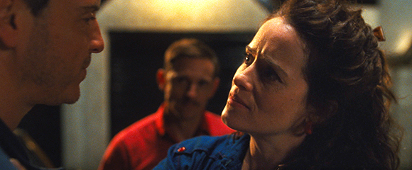 A woman with dark curly hair looks up at a man's profile in a dimly lit room.