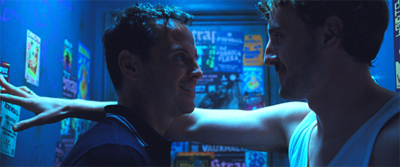 Two men face each other and smile in a room illuminated by blue light, with posters on the wall behind them.
