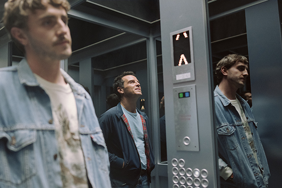Three men stand inside an elevator, one in the foreground and two in the background.