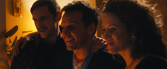 Three smiling adults in a close embrace under warm, dim lighting.