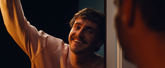A smiling man with short brown hair peeks around a doorframe toward another person.