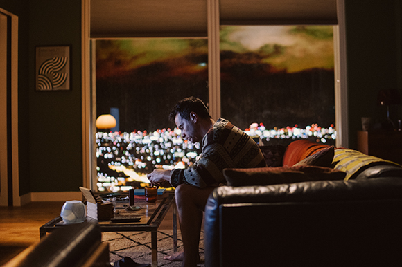 A man sits on a dark leather sofa in a dim room, looking down at a phone with city lights visible through a window.