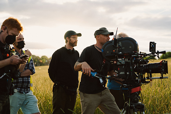 A film crew works with a large professional camera on a tripod in a grassy field during sunset.