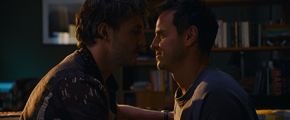 Two men in profile, facing each other closely in a dimly lit room with bookshelves in the background.