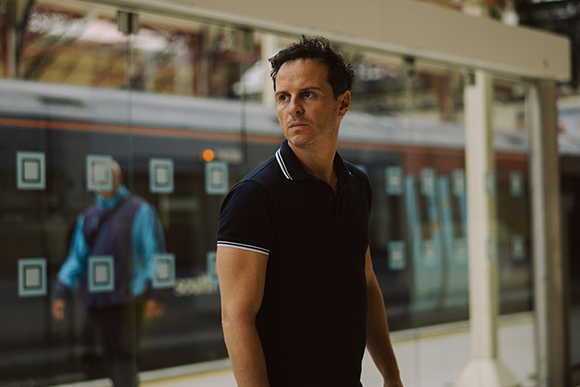 A man in a dark navy polo shirt looks off to the side at a train station platform.