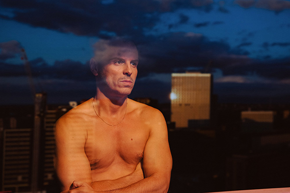 A shirtless man looks off to the side against a dark blue twilight sky with city buildings in the background.