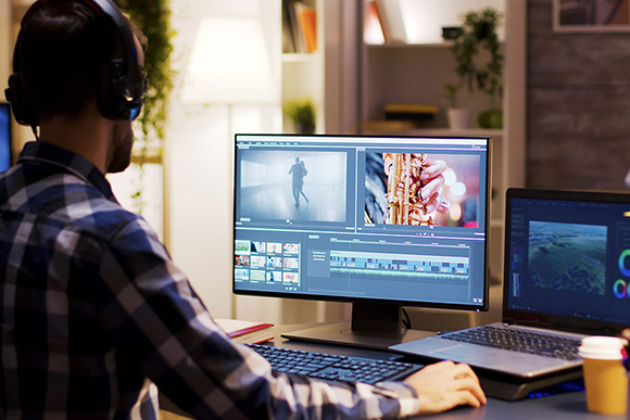 Man wearing headphones works at a desk with a large monitor and a laptop displaying video editing software.