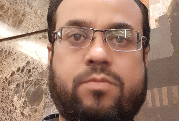 Close-up of a man with dark hair, glasses, and a full beard looking directly at the camera.