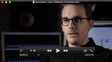 A man with glasses looks forward in a dimly lit studio with audio equipment in the background.