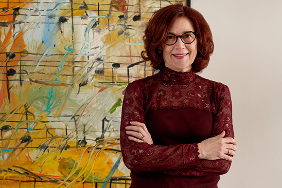 A woman with short red hair and glasses smiles with her arms crossed in front of an abstract musical painting.
