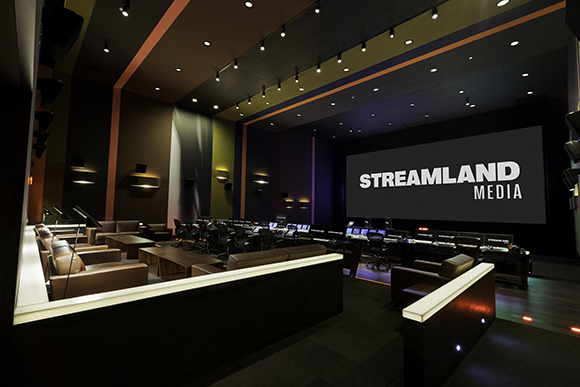 A large dark media studio featuring a massive screen displaying 'STREAMLAND MEDIA' and rows of workstations.