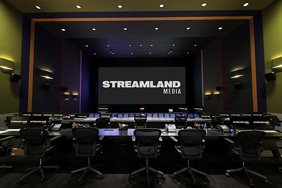 A dark media control room with rows of computer monitors and workstations facing a large screen.