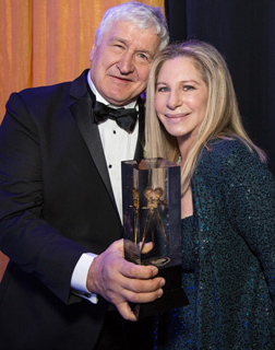 A man in a black tuxedo and a woman with blonde hair hold a tall, clear award trophy together.