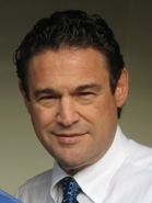 A man with dark curly hair wearing a white collared shirt and a blue patterned tie.