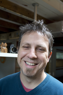 A smiling man with short brown hair and a goatee wearing a blue v-neck shirt.
