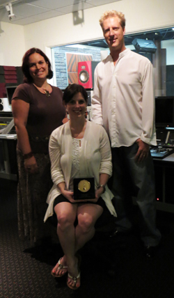 A woman sits holding a gold award while a man and woman stand behind her in a studio.